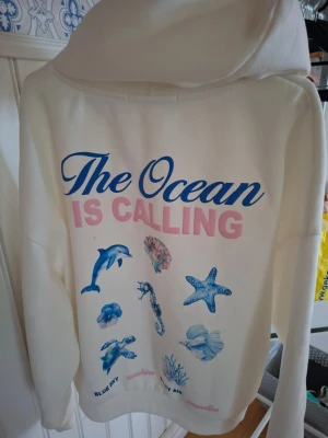 Vit hoodie med havsmotiv och text - Säljer en vit hoodie med stor huva och färgglad print på ryggen. Trycket har texten 'The Ocean is Calling' i blått och rosa samt motiv av delfin, sjöstjärna, snäcka och andra havsdjur. Mjuk och skön, perfekt för dig som gillar strandvibbar och coola prints.