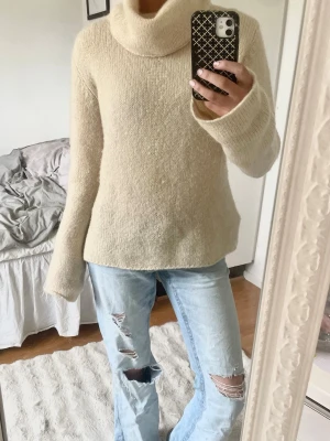 Beige polotröja med vida ärmar i mohair  - Mysig beige stickad polotröja med bred krage och långa, lite vidare ärmar. Tröjan har en rak passform och är gjord i ett mjukt, fluffigt material som känns skönt mot huden. Perfekt för kalla dagar och enkel att styla med jeans eller kjol. Den är cremevit (mellan beige o vit vit)