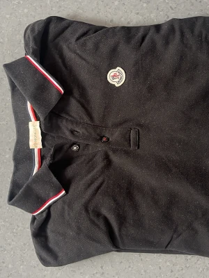 Svart långärmad långärmad piké från Moncler - Snygg svart långärmad skjorta från Moncler med klassisk krage och knappar framtill. Kragen har vita och röda detaljer och bröstet pryds av Monclers ikoniska logga. Perfekt för en clean och stilren look. Storleken är L men passar även M