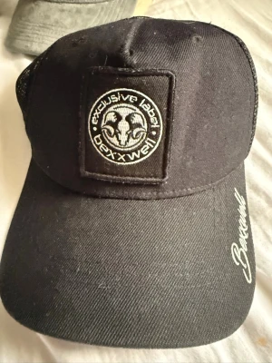Svart keps från Bexxwell - Snygg svart keps från Bexxwell med broderad logga framtill och text på skärmen. Kepsen har mesh på sidorna och bak för extra ventilation och en böjd skärm. Perfekt för en streetwear-look och passar till det mesta.