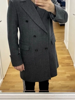 Elegant skräddarsydd dubbelknäpptkappa - Kappan är skräddarsydd på en speciell beställning. Dubbelknäppt, Material: 100% ull 450g med fiskbensmönster i mörkgrå nyans. Kappan är bara prövad. Aldrig använd!