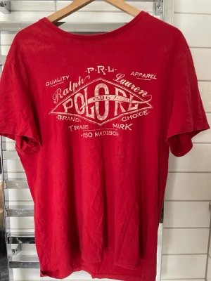 Röd t-shirt från Polo Ralph Lauren - Snygg röd t-shirt från Polo Ralph Lauren med stort vitt tryck framtill. Klassisk rund halsringning och korta ärmar. Tillverkad i mjuk bomull som känns skön mot huden. Perfekt för en avslappnad och stilren look.
