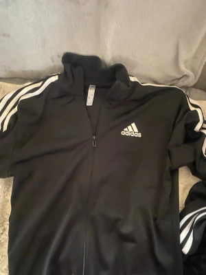 Svart Adidas träningsjacka - Klassisk svart träningsjacka från Adidas med dragkedja framtill och vita ränder längs ärmarna. Jackan har hög krage och Adidas-logga på bröstet. Perfekt för sport eller chill. Materialet är troligtvis polyester.