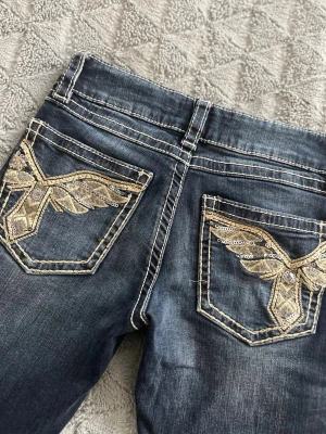Mörkblå jeans med broderade fickor - Snygga mörkblå jeans med unika bakfickor som har guldigt broderi och paljettdetaljer i form av vingar. Jeansen har kontrastsömmar och klassisk femficksmodell. Perfekta för dig som vill sticka ut med coola detaljer.