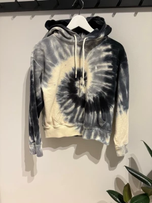 Svart och vit tie-dye hoodie - Cool hoodie med svart och vit tie-dye spiral på både fram- och baksidan. Tröjan har huva med vita snören och ribbade muddar. Materialet ser ut att vara mjukt bomullstyg, perfekt för en avslappnad och trendig look.