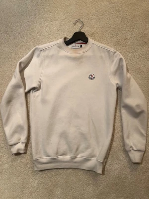 Beige sweatshirt från Moncler - Säljer en stilren beige sweatshirt från Moncler med rund halsringning och diskret Moncler-logga broderad på bröstet. Tröjan har ribbade muddar vid ärmslut och nederkant. Perfekt för en clean och avslappnad look.
