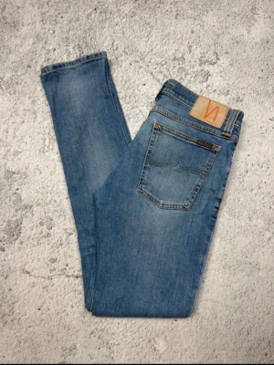 Blå slim fit jeans från Nudie Jeans - Säljer ett par ljusblå slim fit jeans från Nudie Jeans med klassisk femficksmodell och snygga slitningar. Jeansen har en skinnpatch med logga bak och är tillverkade i mjuk denim. Perfekta för en avslappnad och trendig look.  W30 L34