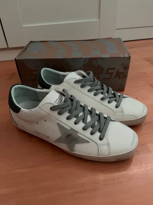 Golden Goose vita sneakers med grå stjärna - Snygga vita sneakers från Golden Goose med grå stjärna på sidan, grå skosnören och svart detalj på hälen. Skorna har en klassisk låg silhuett och är tillverkade i läder med slitna detaljer för en cool look. Perfekta för dig som gillar streetstyle.