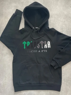 Trapstar hoodie - Svart hoodie med broderad text i grönt, vitt och grått på bröstet. Klassisk känguruficka framtill, justerbar huva med snören och ribbade muddar. Tillverkad i mjukt bomullsmaterial för en skön och avslappnad look.