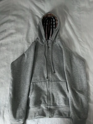 Grå zip hoodie från Burberry - Snygg grå zip hoodie från Burberry med klassiskt rutigt foder i huvan och dragsko. Hoodien har två Pockets framtill och ribbade muddar. Perfekt för dig som vill ha en stilren och exklusiv look med en twist av Burberrybränd design.