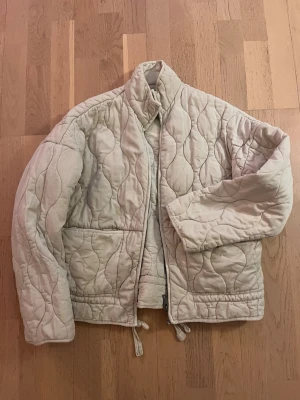 Beige quiltad pufferjacka från Zara - Säljer en gräddvit quiltad jacka från Zara med hög krage och dragkedja. Jackan har två stora fickor framtill och justerbar dragsko i nederkant. Materialet är mjukt och vadderat. Enkel men trendig design som passar till det mesta.