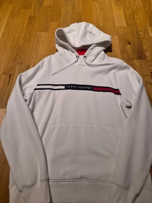 Vit hoodie från Tommy Hilfiger - Snygg vit hoodie från Tommy Hilfiger med klassisk logga och ränder i svart och rött över bröstet.  Använd en gång så den är absolut topp skick 