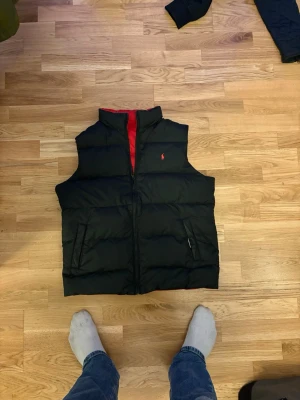 Vändbar dunväst från Polo Ralph Lauren - Snygg vändbar dunväst från Polo Ralph Lauren med ena sidan svart och andra sidan röd. Västen har hög krage, dragkedja framtill och två fickor på varje sida. Klassisk liten logga på bröstet. Perfekt för lager-på-lager och streetwear-vibe.
