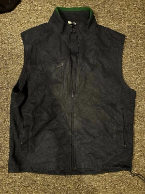 Mörkblå väst från Polo Ralph Lauren - Stilren mörkblå väst från Polo Ralph Lauren med dragkedja framtill, hög krage och diskret broderad logga på bröstet. Västen har fickor med dragkedja och är tillverkad i ett mjukt, mockaliknande material. Perfekt för lager på lager och en clean look.