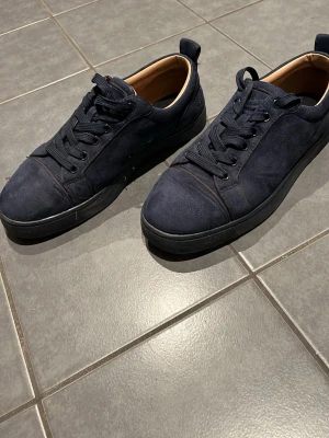 Mörkblå sneakers från Christian Louboutin - Snygga mörkblå sneakers i mocka från Christian Louboutin med klassisk röd sula och svarta skosnören. Lågt skaft och stilren design som passar till det mesta. Ikonisk signatur på sulan. Den har en tunn sula där inne.