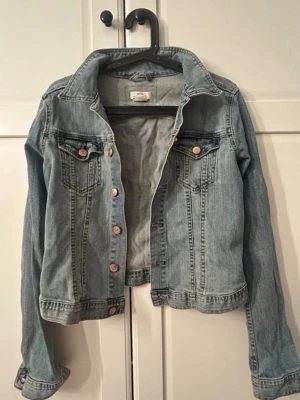 Ljusblå jeansjacka från &NOW - Ljusblå jeansjacka från &NOW med klassisk krage, bröstfickor med knapp och snygga kopparfärgade knappar framtill.