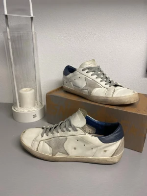 Golden Goose vita sneakers med blå detalj - Stilrena vita sneakers från Golden Goose med grå stjärndetalj på sidan, grå skosnören och blå hälkappa. Ovandel i läder och mocka, klassisk låg modell med vintage-inspirerad look. Perfekta för dig som gillar en avslappnad och trendig stil.