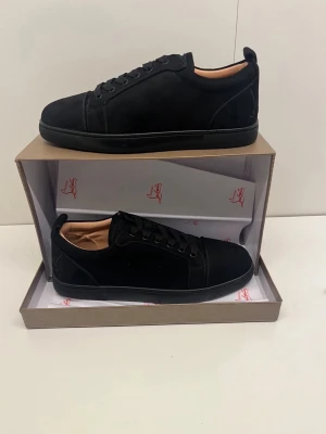 Svarta sneakers från Christian Louboutin - Stilrena svarta sneakers från Christian Louboutin i mocka med svarta skosnören och diskret logga på hälen. Låg modell med svart sula och beige innerfoder. Perfekta för dig som gillar en clean och lyxig look.