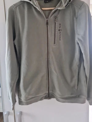 Grå hoodie från Sail Racing - Snygg grå hoodie från Sail Racing med dragkedja framtill och en bröstficka med zip. Hoodien har en broderad logga på bröstet och två sidofickor. Tillverkad i mjukt material som känns skönt mot huden. Perfekt för en avslappnad stil.