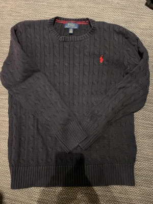 Mörkblå kabelstickad tröja Polo Ralph Lauren - Kabelstickad mörkblå tröja från Polo Ralph Lauren med klassisk röd broderad logga på bröstet. Tröjan har rund halsringning, ribbade muddar och är långärmad. Perfekt för dig som gillar stilrena och tidlösa plagg. Det är bara att höra av er vid minsta lilla fråga! Priset är inte hugget i sten!