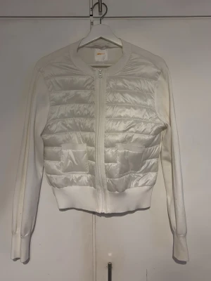 Vit quiltad cardiganjacka - Snygg vit cardiganjacka från GinaTricot med quiltad framsida i glansigt material och stickade ärmar samt rygg. Jackan har dragkedja framtill och ribbade muddar. Perfekt för lager-på-lager och en clean look.