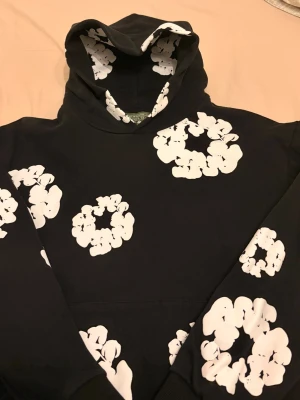 Svart hoodie från Denim Tears - Svart hoodie från Denim Tears med stora vita blomliknande tryck över hela plagget. Klassisk huva och magficka framtill. Skön och avslappnad passform, perfekt för dig som vill sticka ut med en unik design. Materialet känns mjukt och bekvämt.