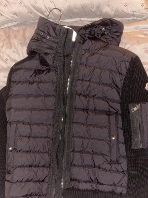 Svart pufferjacka med stickade ärmar - Snygg svart pufferjacka med stickade ärmar och huva. Jackan har dragkedja framtill, två fickor med tryckknappar och en extra ficka på ärmen. Materialet är en mix av quiltad nylon och ribbstickad bomull, perfekt för kalla dagar.