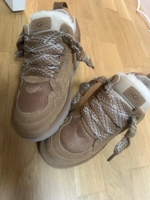UGG bruna sneakers med fårskinn - Snygga bruna sneakers från UGG med grova snören i beige och vitt. Ovandelen är i mocka och mesh, och insidan är fodrad med mjukt vitt fårskinn. Skorna har en chunky sula och UGG-logga både fram och bak. Perfekta för dig som vill ha både stil och komfort.