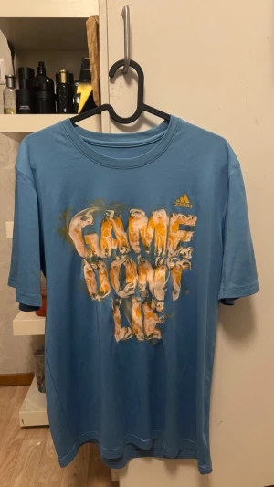 Blå Adidas t-shirt med tryck - Säljer en blå t-shirt från Adidas med coolt tryck på bröstet där det står 'GAME DON'T LIE' i orange och vitt. T-shirten har korta ärmar, rund hals och är gjord i ett mjukt material som känns skönt mot huden. Perfekt för dig som gillar sportig stil. Säg till om du behöver fler bilder 🙌