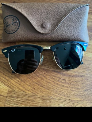 Ray-Ban Clubmaster svarta solglasögon - Säljer ett par klassiska Ray-Ban Clubmaster solglasögon med svart ovandel och guldfärgade detaljer. Glasen är mörka och bågarna har en retroinspirerad form. Kommer med original Ray-Ban fodral i brunt konstläder. ÄKTA!!!