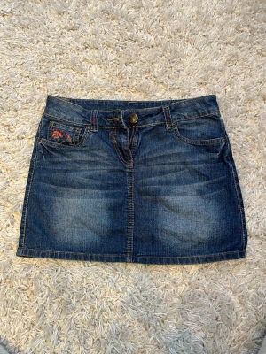 Jeanskjol - Snygg jeanskjol med låg midja och en fin detalj på ena framfickan ett litet blombrodderi 🌹 Midjemått rakt över 35 cm & kjolen är ca 33 cm lång💕