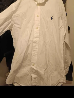 Vit skjorta från Ralph Lauren - Klassisk vit skjorta från Ralph Lauren med blå logga på bröstet. Slim fit-modell i 100% bomull med stretch för extra komfort. Skjortan har långa ärmar och knappar framtill. Perfekt till en clean och stilren look.