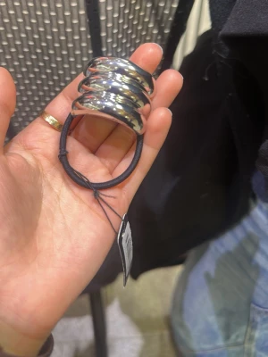 Silvrig hårsnodd från Montini - Snygg hårsnodd från Montini med en chunky, silvrig metallring som dekoration. Elastiskt svart band som håller håret på plats och ger en cool touch till din look. Perfekt accessoar för dig som vill ha något extra i håret.