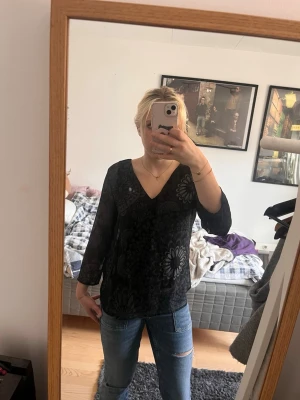 Svart blommig blus med V-ringning Desigual  - Snygg svart blus med stora blommönster och V-ringad hals. Blusen har trekvartsärm och är tillverkad i ett tunt, lätt material som ger en avslappnad men stilren look. Perfekt att matcha med jeans eller kjol för en trendig outfit.