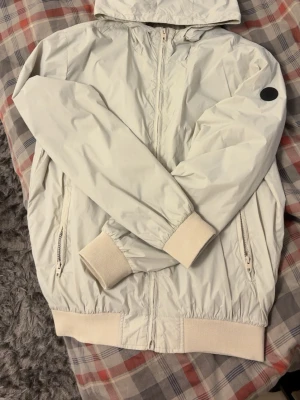 Vit vindjacka med huva - Stilren vit vindjacka med huva och dragkedja framtill. Jackan har ribbade muddar vid ärmslut och nederkant samt två fickor med dragkedja. En svart patch-detalj på ena ärmen ger en cool touch. Perfekt för blåsiga dagar.