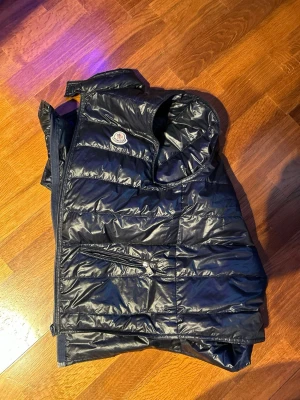 Mörkblå quiltad dunväst från Moncler - Snygg mörkblå dunväst från Moncler med quiltad design och hög krage. Västen har dragkedja framtill, två fickor med dragkedja och klassisk Moncler-logga på bröstet. Perfekt för lager-på-lager och riktigt stilren.