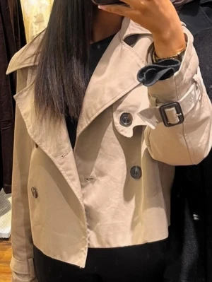 Kort beige trenchcoat med bälte - Trendig kort trenchcoat i beige med dubbelknäppning och stora knappar. Jackan har bred krage, axelklaffar och ett bälte med spänne vid ärmsluten för extra detalj. Perfekt för en stilren och modern look.