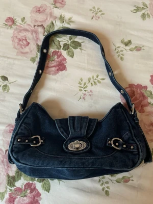 Mörkblå denim handväska med spännen - Trendig mörkblå handväska i denim med silverfärgade detaljer och spännen på framsidan. Väskan har ett vridlås och justerbart axelband med nitar. Perfekt accessoar för en cool och avslappnad stil.