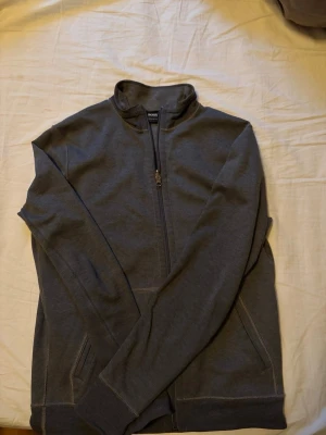 Mörkgrå zip hoodie från Hugo Boss - Snygg mörkgrå hoodie från Hugo Boss med dragkedja framtill och regular fit. Tröjan har hög krage, långa ärmar och två fickor.