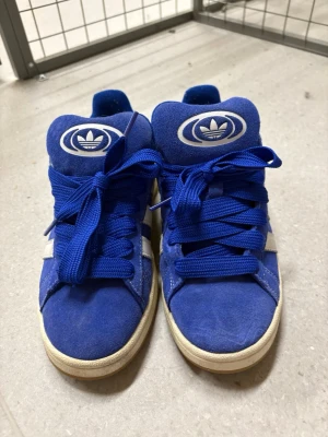 Blå Adidas sneakers med mocka - Snygga blå sneakers från Adidas med klassiska vita ränder på sidan och stort Adidas-logga på plösen. Skorna är gjorda i mocka och har tjocka blå skosnören samt vit sula. Perfekta för dig som gillar retrostil och streetwear.