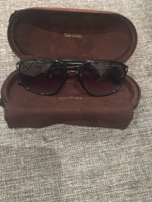 Tom Ford solglasögon  - Snygga mörkblå aviator solglasögon från Tom Ford med mörka glas och tunna metallbågar. Kommer i originalbrunt fodral med Tom Ford-logga. Perfekta för dig som vill ha en klassisk och stilren look.