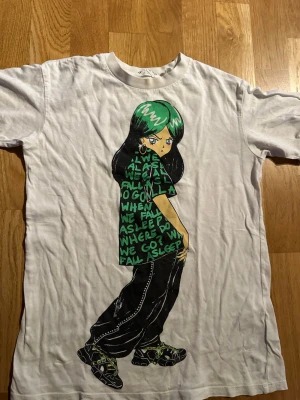 Vit t-shirt med Billie Eilish tryck från H&M - Säljer en vit t-shirt från H&M med ett Billie Eilish tryck och text på framsidan. T-shirten har rund hals och är tillverkad i mjuk bomull. Perfekt för dig som gillar streetwear och unika motiv.