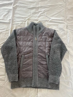 Grå stickad cardigan med quiltad front - Snygg grå cardiganjacka med stickade ärmar och rygg samt quiltad front i blankt material. Jackan har hög krage, dragkedja och två fickor fram. Perfekt för lager på lager och ger en modern look till din outfit.