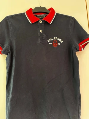 Marinblå piké från Sail Racing - Klassisk marinblå pikétröja från Sail Racing med röd krage och ärmslut, samt vit broderad logga och detaljer på bröstet. Tröjan har korta ärmar och knappar vid halsen. Perfekt för en sportig och stilren look.