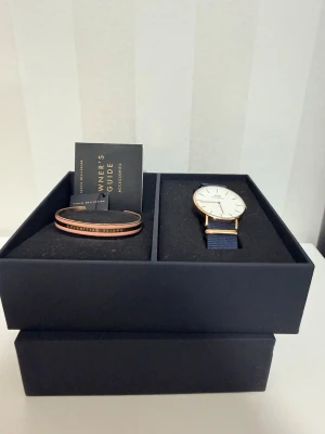 Daniel Wellington klockset blå/guld - Snyggt klockset från Daniel Wellington med en klassisk klocka i guld och vitt, samt blått tygarmband. Medföljer matchande guldarmband med gravyr. Perfekt accessoar för dig som gillar stilrena detaljer.