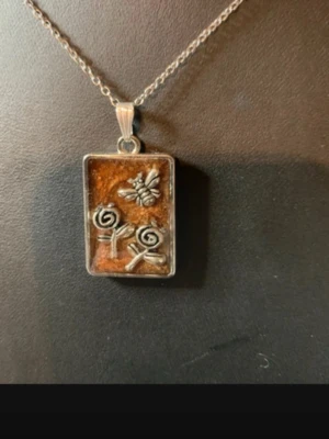 Halsband med rektangulärt hänge och bi - Unikt halsband med ett rektangulärt hänge i silver färg. Hänget har en glittrande brun bakgrund och dekoreras med en liten bi och två stiliserade blommor i silverfärg. Kedjan är tunn och silverfärgad. Perfekt för dig som gillar naturinspirerade smycken.