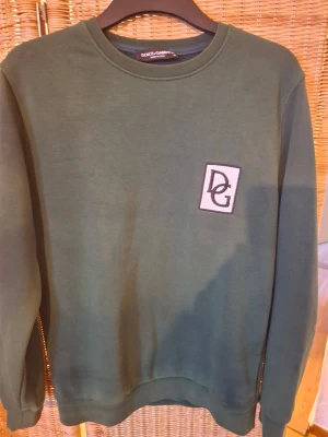 Grön sweatshirt från Dolce & Gabbana - Snygg grön sweatshirt från Dolce & Gabbana med DG-logga på bröstet. Tröjan har rund halsringning och ribbade muddar vid ärmslut och nederkant. Tillverkad i mjukt material som känns skönt mot huden. Perfekt för en avslappnad och trendig look.