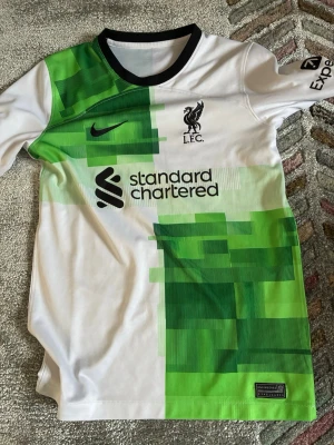 Liverpool FC bortatröja Nike grön/vit - Säljer en Liverpool FC bortatröja från Nike i vitt och grönt med digitalt pixelmönster. Tröjan har svart krage, klubbmärke och sponsortryck på bröstet samt Nike-logga. Tillverkad i lätt och ventilerande material, perfekt för fotbollsträning eller match.