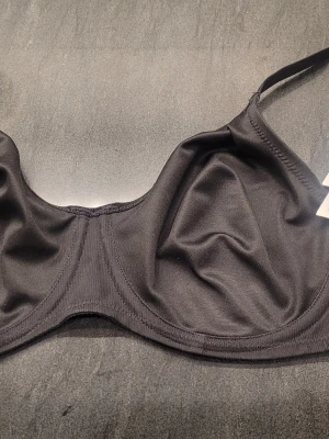 Svart bygel-bh från Triumph 75 D - Snygg svart bygel-bh från Triumph, modell Urban Minimizer W. Tillverkad i mjukt material med 49% polyester, 33% polyamid och 18% elastan.Ger en slät siluett och justerbara axelband. Mycket bekväm.