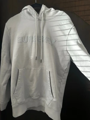 Ljusblå hoodie från Burberry - Snygg ljusblå hoodie från Burberry med stor logga i relief på bröstet. Hoodien har dragsko i huvan och en klassisk magficka. Tillverkad i mjukt bomullsmaterial som känns skönt mot huden. Perfekt för en clean och stilren look.